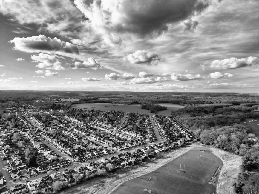 An aerial black-and-white photograph showing a densely populated residential area with rows of houses arranged along streets, surrounding a sports field with visible goalposts and a running track. The houses have uniform designs with pitched roofs, and are closely packed with small gardens and driveways. To the right, there is a wooded area with trees and open fields extending to the horizon. Above, the sky features a mix of large, billowing clouds and clear patches, casting shadows that add depth to the scene. The landscape appears flat, with the residential zone bordered by greenery and open land, suggesting a suburban setting. Such environments may benefit from private rubbish collection services like those offered by House Clearance Watford, especially for waste generated during home or garden clearance. The scene indicates a typical suburban environment where on-site or independent rubbish removal could be an alternative to local authority collections, particularly in managing bulk waste or large quantities of refuse from residential or community areas. This comprehensive view underscores the importance of effective waste management solutions in accommodating the needs of such communities.