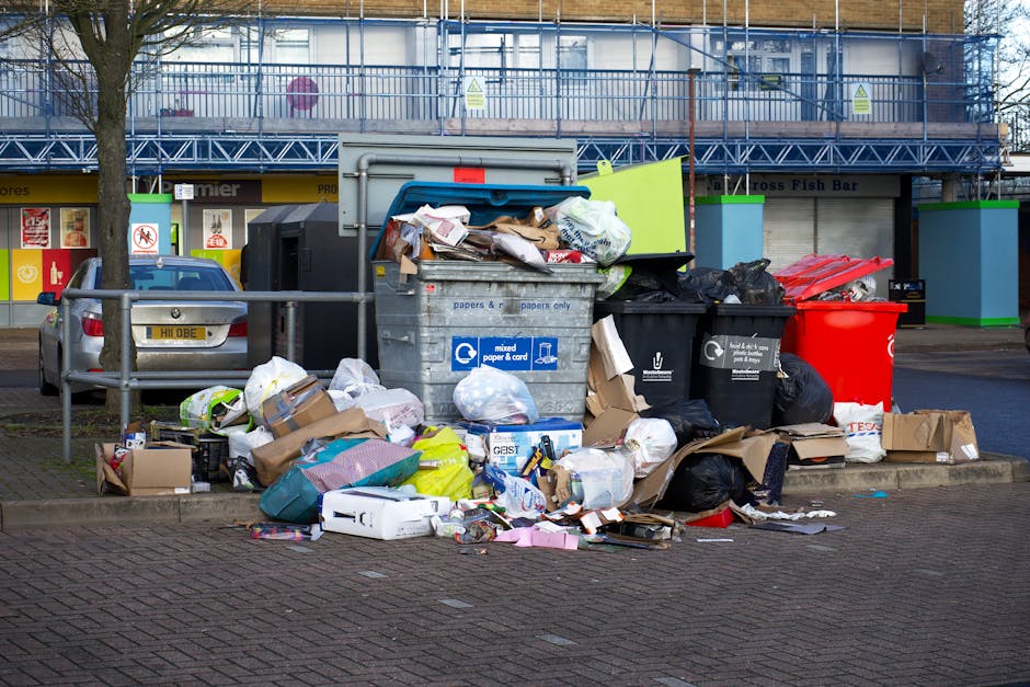 A large collection of overflowing rubbish bins and piles of discarded household waste and packaging materials are situated on a paved sidewalk area, partially surrounded by a metal barrier. The collection includes various black, green, and red plastic wheelie bins with lids left open, revealing accumulated waste such as cardboard boxes, plastic bags filled with general rubbish, paper and paperboard cartons, and miscellaneous packaging debris. The waste spills onto the surrounding pavement and street, with some items like flattened cardboard and plastic containers extending outward. Behind the rubbish, a silver car with a yellow registration plate is parked on a neighboring parking space. In the background, there are commercial storefronts with signage, and a multi-storey building under renovation or construction, covered in blue scaffolding and safety netting. The scene is lit by ambient daylight, highlighting the untidy state of the waste accumulation, which may suggest an issue needing a private or professional rubbish removal service like those provided by House Clearance Watford, especially for on-site clearance or alternative waste handling outside standard municipal collection.