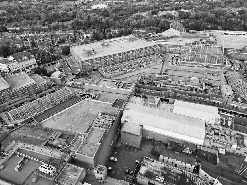An aerial black-and-white photograph showing a densely populated residential area with rows of houses arranged along streets, surrounding a sports field with visible goalposts and a running track. The houses have uniform designs with pitched roofs, and are closely packed with small gardens and driveways. To the right, there is a wooded area with trees and open fields extending to the horizon. Above, the sky features a mix of large, billowing clouds and clear patches, casting shadows that add depth to the scene. The landscape appears flat, with the residential zone bordered by greenery and open land, suggesting a suburban setting. Such environments may benefit from private rubbish collection services like those offered by House Clearance Watford, especially for waste generated during home or garden clearance. The scene indicates a typical suburban environment where on-site or independent rubbish removal could be an alternative to local authority collections, particularly in managing bulk waste or large quantities of refuse from residential or community areas. This comprehensive view underscores the importance of effective waste management solutions in accommodating the needs of such communities.