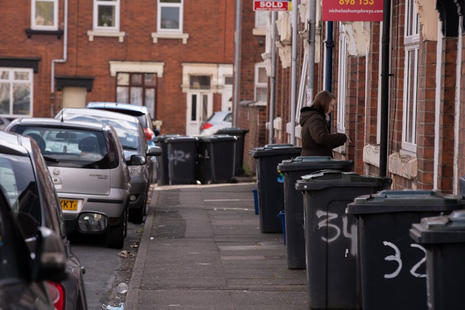 A street scene showing a row of dark green and black wheelie bins lined up along the pavement outside brick residential buildings with white-framed windows. The bins are positioned in front of the houses, some with their lids closed, and have white graffiti markings visible on their sides. A woman standing nearby is dressed in a brown jacket and blue jeans, appearing to be opening or retrieving items from one of the bins. In the background, additional bins are placed against the building facade, with parked cars visible on the road, including a silver hatchback and other vehicles further along the street. The environment suggests a typical urban residential area, likely undergoing or preparing for rubbish clearance, with the scene emphasizing private waste management outside homes as part of an independent rubbish removal service. The lighting indicates daytime with natural light illuminating the scene, and the overall atmosphere reflects routine household waste disposal activities occurring on a residential street.