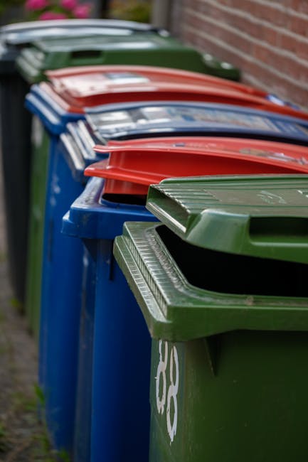 A row of large, plastic wheelie bins lined up outdoors along a paved pathway adjacent to a brick wall. The bins are predominantly blue, with green and red lids, arranged in an alternating pattern. The green lids are slightly raised, revealing the openings, while the red lids are closed on top. The surface of the bins shows slight texture and wear, with some visible scratches and marks. The background features a blurred view of a residential area, with green foliage and an overcast sky. This outdoor setting illustrates typical arrangements for household waste disposal that could be associated with private rubbish collection or small-scale waste handling, as managed by companies like House Clearance Watford, rather than municipal curbside collection.
