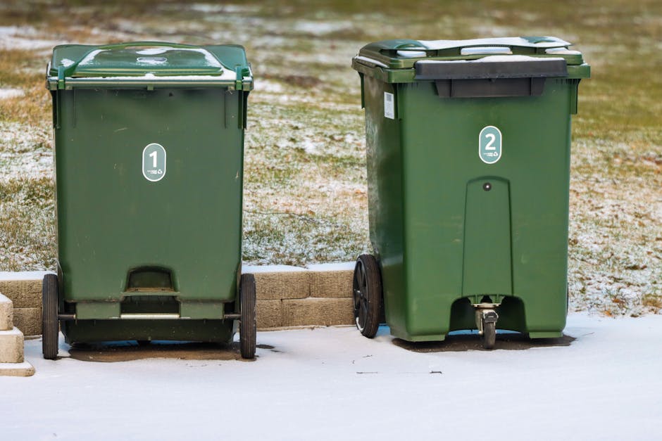 Two green wheeled rubbish bins are positioned side by side on a concrete surface, with the left bin marked with the number '1' and the right bin marked with the number '2'. The bins are made of durable plastic with textured surfaces; the left bin has a slightly shiny finish with minor scratches, while the right bin appears matte with a black lid. The left bin’s lid has a white latch, and the right bin’s lid features a black handle strip and a secured black lid latch. Both bins are placed on a paved area adjacent to a low brick border, with a patchy, snow-dusted ground in the background and a grassy yard beyond. The lighting is natural, suggesting daytime with overcast conditions, creating a soft illumination across the scene. The environment indicates typical outdoor waste collection points, supporting the context of private rubbish handling and waste disposal or collection options separate from local council services, consistent with alternative rubbish removal practices. The overall visual emphasizes the practical aspect of waste management and on-site clearance resources provided by companies like House Clearance Watford.